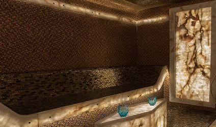 Interior de una lujosa sala de vapor con paredes de mosaico dorado, bancos integrados y paneles de piedra natural retroiluminados.