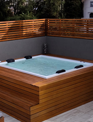 Bañera de hidromasaje cuadrada instalada en una terraza, rodeada por una estructura y valla de madera de diseño moderno.