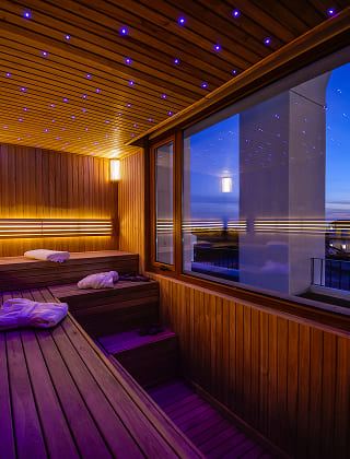 Interior de una sauna de madera con iluminación cenital con efecto cielo estrellado y un gran ventanal al atardecer.