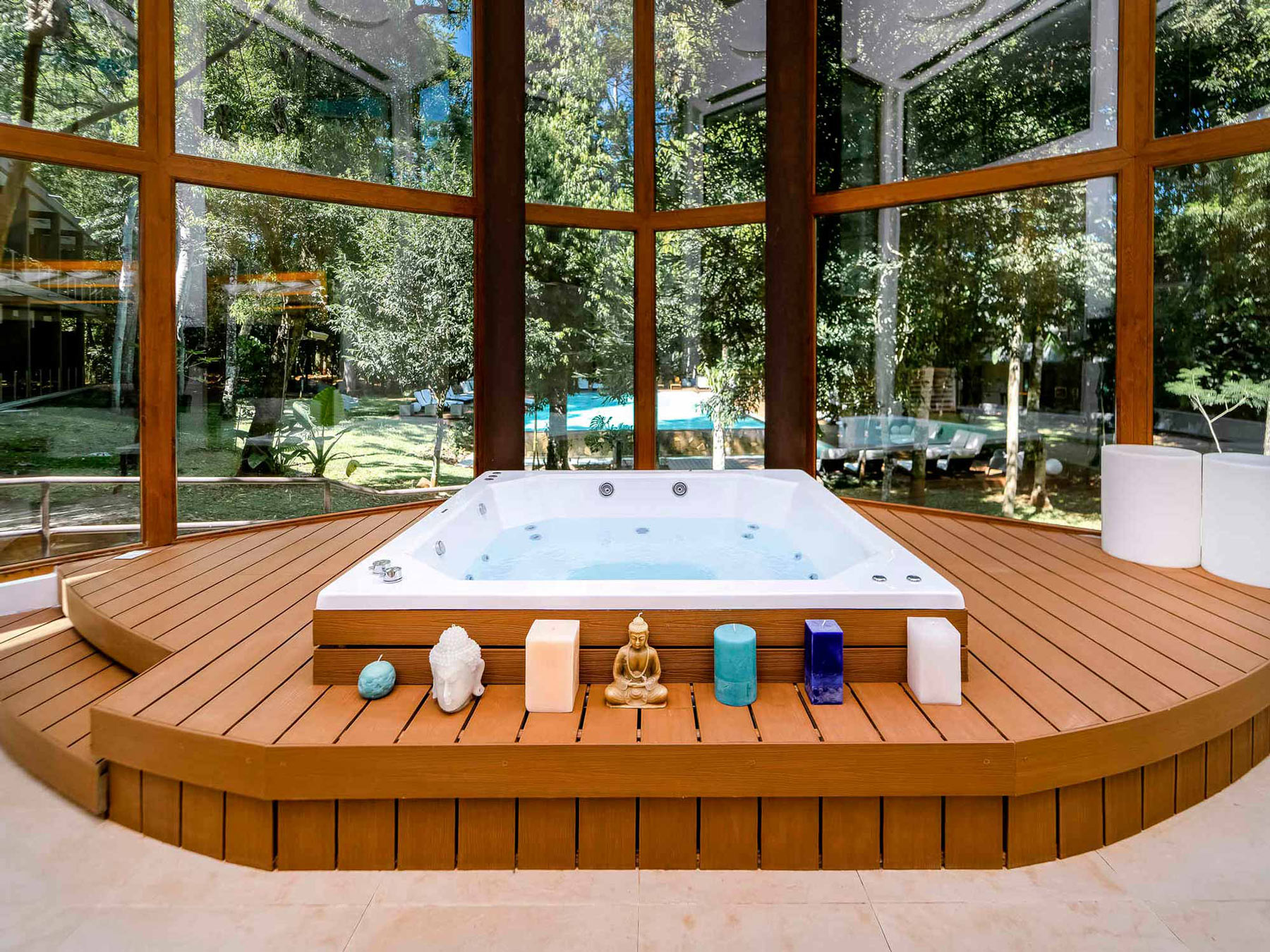 Bañera de hidromasaje cuadrada integrada en un deck de madera dentro de un gazebo acristalado con vistas a un jardín, decorada con velas y figuras zen.